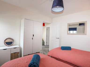A bedroom with two single beds with coral bedspreads and blue towels a white wardrobe a white dresser with a round mirror and a wall mirror at 19 Parc Delfryn in Benllech