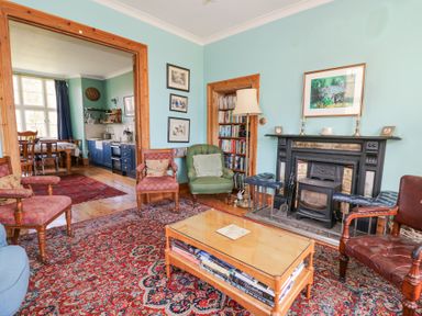 A living room with a fireplace, armchairs, a coffee table with books, bookshelf, and an adjoining kitchen at Plas Pontfaen near Fishguard and Newport Pembrokeshire