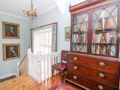 A stair landing with a wooden bookcase filled with books a brown leather chair and framed portraits and a window with curtains at Plas Pontfaen near Fishguard and Newport Pembrokeshire