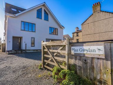 A white house with multiple windows and a wooden gate with a sign that says Plas Gwylan in Rhosneigr