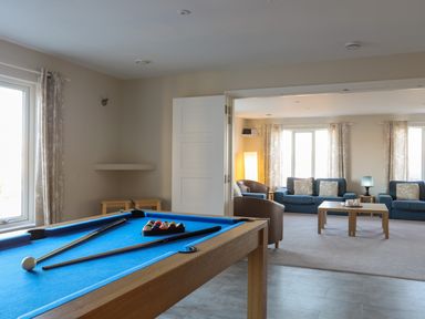 A room with a blue pool table and two pool cues a white ball and a rack of balls next to a living area with blue sofas brown chairs a wooden coffee table and windows with curtains at Plas Gwylan in Rhosneigr