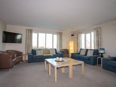 A living room with blue sofas brown armchairs a wooden coffee table and a wall-mounted television at Plas Gwylan in Rhosneigr