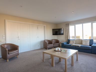 A living room with brown armchairs a blue sofa a wooden coffee table and a wall-mounted TV at Plas Gwylan in Rhosneigr