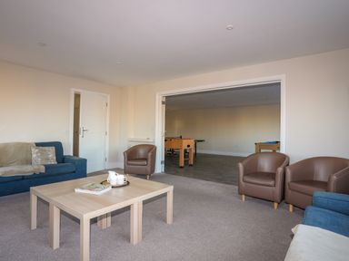 A living room with blue sofas brown armchairs and a wooden coffee table opening to a games room at Plas Gwylan in Rhosneigr