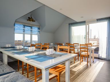 A dining area with white tables wooden chairs blue placemats and mugs by a window with blue curtains at Plas Gwylan in Rhosneigr
