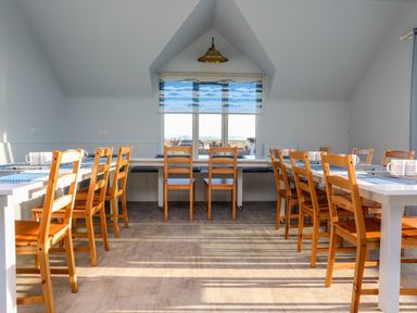 A dining room with wooden chairs around white tables and a window with blue patterned blinds at Plas Gwylan in Rhosneigr