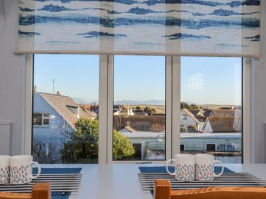 A window with a blue patterned blind overlooking rooftops and distant hills with mugs on a table at Plas Gwylan in Rhosneigr
