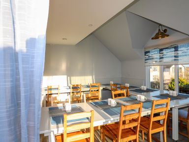 A dining room with wooden chairs and white tables set with placemats and mugs at Plas Gwylan in Rhosneigr