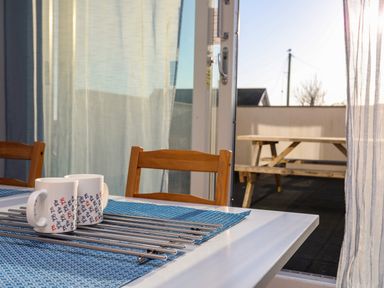 A dining table with two mugs and metal skewers on a blue mat next to wooden chairs and an open door showing an outdoor wooden picnic table at Plas Gwylan in Rhosneigr