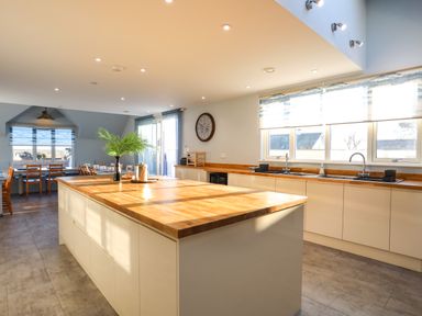 A kitchen with a large wooden island countertop and dining tables with chairs in the background at Plas Gwylan in Rhosneigr