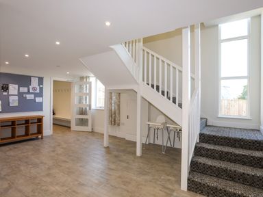 A hallway with stairs carpeted leading up and down two high chairs under the stairs with a wooden shelf and notice board on the wall at Plas Gwylan in Rhosneigr