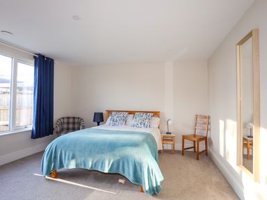 A bedroom with a bed covered with a blue blanket two pillows two bedside tables lamps a chair a mirror and blue curtains at Plas Gwylan in Rhosneigr