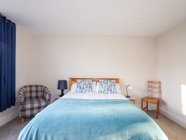 A bedroom with a bed with blue and white pillows a blue blanket two bedside tables with lamps a plaid armchair and a wooden chair at Plas Gwylan in Rhosneigr