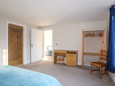 A bedroom with a blue bedspread a wooden desk and stool a wooden chair and an open walk-in shower at Plas Gwylan in Rhosneigr