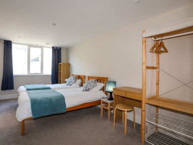 A bedroom with two single beds a desk a stool a lamp a small open wardrobe and a window with blue curtains at Plas Gwylan in Rhosneigr