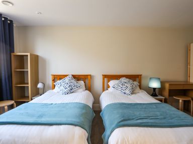 A bedroom with two single beds wooden headboards blue blankets white pillows patterned cushions wooden shelves desks and lamps at Plas Gwylan in Rhosneigr