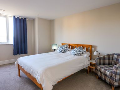 A bedroom with a bed with white bedding and patterned pillows a plaid armchair and a window with blue curtains at Plas Gwylan in Rhosneigr