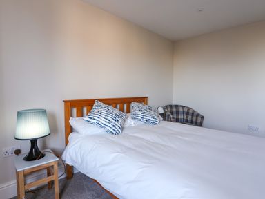 A bedroom with a wooden bed white bedding blue patterned pillows a bedside table with a lamp and a plaid upholstered armchair at Plas Gwylan in Rhosneigr