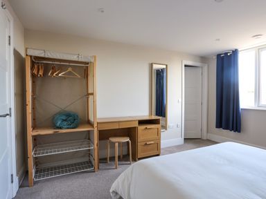 A bedroom with an open wardrobe with hangers a wooden desk with a stool a wall mirror blue curtains and a bed at Plas Gwylan in Rhosneigr