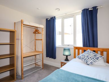 A bedroom with a wooden bed a side table with a lamp blue curtains an open wardrobe with hangers and a wooden bookshelf at Plas Gwylan in Rhosneigr