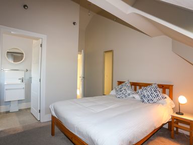 A bedroom with a wooden bed with white bedding and two pillows a small side table with a lamp and an open door leading to a bathroom at Plas Gwylan in Rhosneigr