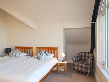 A bedroom with two single beds a nightstand with a lamp a plaid armchair and a window with dark curtains at Plas Gwylan in Rhosneigr