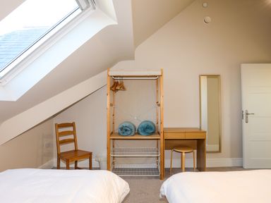 A bedroom with two beds a wooden chair a wooden shelving unit with hangers and blankets a wooden desk with a stool a wall mirror and a skylight window at Plas Gwylan in Rhosneigr
