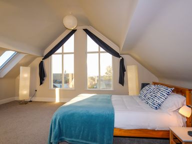 A bedroom with a wooden bed with blue and white pillows and a blue blanket under a sloped ceiling with large triangular windows and two tall floor lamps at Plas Gwylan in Rhosneigr