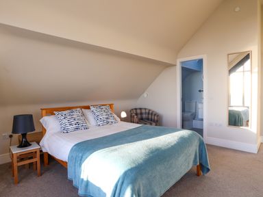 A bedroom with a wooden bed with blue and white pillows and a blue blanket a black lamp on a wooden stool a plaid armchair a wall mirror and an open door to a bathroom at Plas Gwylan in Rhosneigr