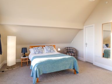 A bedroom with a wooden bed with blue and white pillows a blue blanket a tall white floor lamp a small black bedside lamp a plaid armchair a door and a wall mirror at Plas Gwylan in Rhosneigr