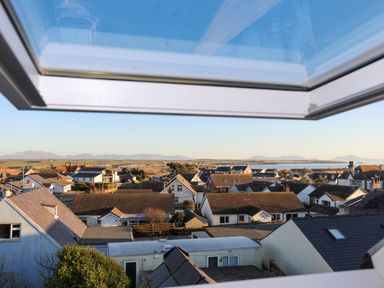 A view of rooftops and houses with mountains and sea in the distance seen through an open skylight window at Plas Gwylan in Rhosneigr