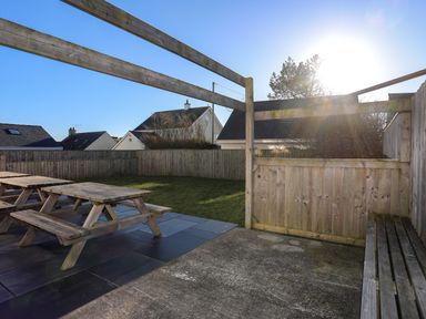 An outdoor area with wooden picnic tables, a tiled patio, grass, and wooden fence at Plas Gwylan in Rhosneigr