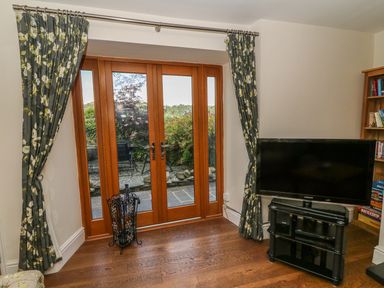A room with wooden double doors leading outside floral curtains a television on a black stand and a bookshelf