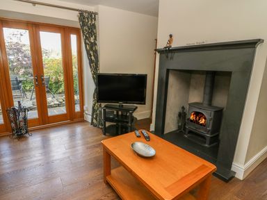 A living room with a wood stove fireplace television on a stand wooden coffee table with remotes and glass doors to a garden at Woodbine Cottage in Ambleside