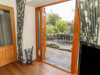 A view from inside a room with wooden floor and floral curtains through open wooden framed glass doors to an outdoor patio with stone tiles and garden furniture at Woodbine Cottage in Ambleside