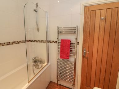 A bathroom with a bathtub shower combination a glass shower screen a heated towel rail with a red towel and a wooden door at Woodbine Cottage in Ambleside