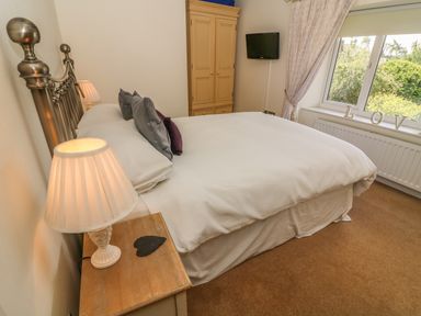 A bedroom with a double bed a wooden bedside table a lamp a wardrobe a wall mounted television and a window with curtains at Woodbine Cottage in Ambleside