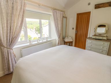 A bedroom with a bed covered in white bedding a window with curtains a dresser with a mirror a wooden door and a mirror on the wall at Woodbine Cottage in Ambleside
