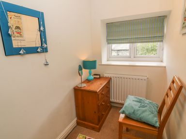 A small room with a wooden chair and cushion next to a wooden chest of drawers with a lamp and mirror near a window with striped blinds at Woodbine Cottage in Ambleside