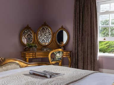 A bedroom with a dressing table with three mirrors a chair with patterned upholstery a bed with a folded magazine and a window with curtains at Hunters Moon in Charlestown