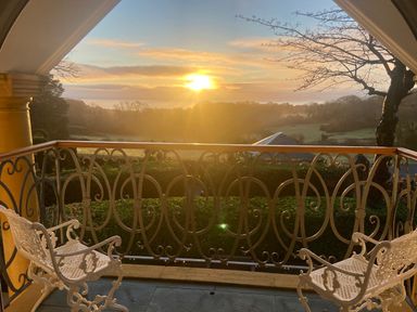 A balcony with two white chairs and a decorative railing overlooking trees and fields at sunrise at Hunters Moon in Charlestown
