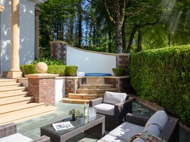 An outdoor seating area with wicker furniture and a glass table near steps leading to a hot tub surrounded by greenery at Hunters Moon in Charlestown