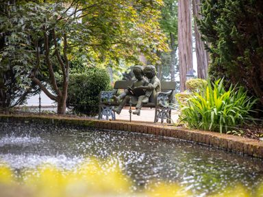 A garden pond with a statue of two children reading on a bench surrounded by trees and plants at Hunters Moon in Charlestown