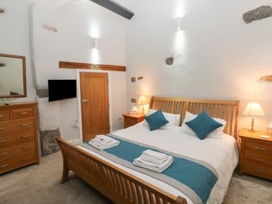 A bedroom with a wooden bed frame blue cushions white linens bedside tables with lamps a wooden dresser and a wall-mounted television at Blacksmiths Cottage Pembroke