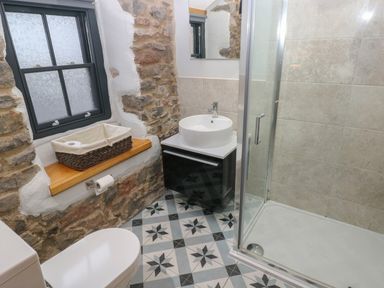 A bathroom with a toilet, sink with vanity, shower enclosure, patterned floor tiles, stone wall, and window at Blacksmiths Cottage in Pembroke