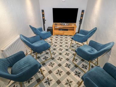 A small room with six blue chairs surrounding a television on a wooden stand with two tall speakers at Blacksmiths Cottage in Pembroke
