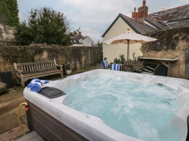 An outdoor patio area with a bubbling hot tub wooden bench table with chairs and umbrella at Blacksmiths Cottage in Pembroke