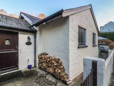 An exterior view of a white stone cottage with stacked firewood next to a dark wooden door at Old Farm Cottage Woolacombe