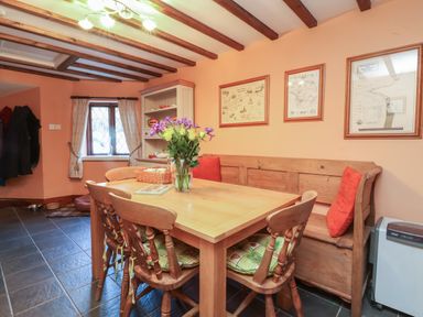 A dining area with a wooden table and chairs with cushions a corner bench with cushions a vase with flowers on the table maps on the wall and a window at Old Farm Cottage in Woolacombe