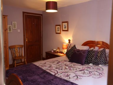 A bedroom with a wooden bed and multiple pillows a nightstand with a lamp and decorations a wooden chair and wardrobe a door on a purple wall at Old Farm Cottage in Woolacombe